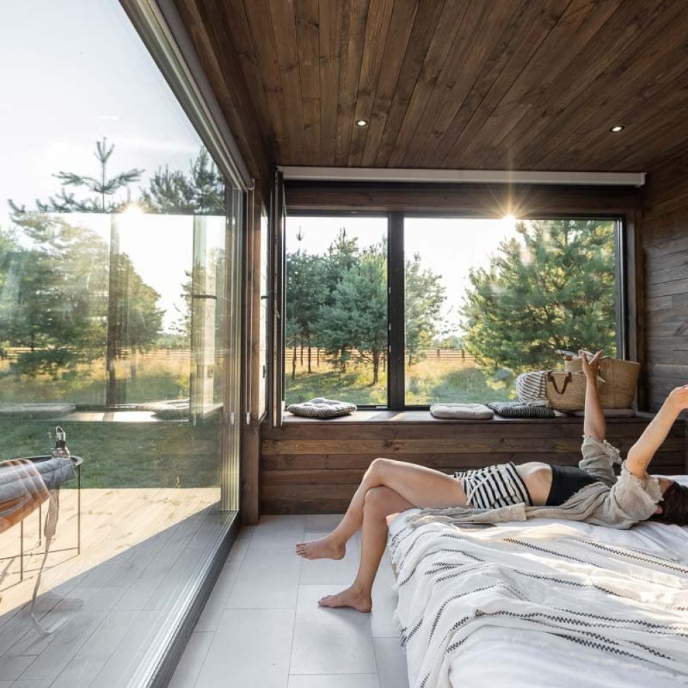 Woman wakes up in a country house or hotel with panoramic windows in pine forest lying on the bed and raised her hands. Good morning and recreation on nature concept